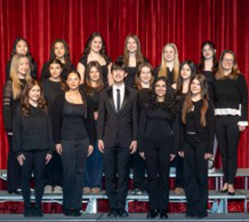 Group portrait of a high school senior dance ensemble standing on tiered risers in front of a red stage curtain, dressed in coordinated black performance attire and posing for a formal photo.